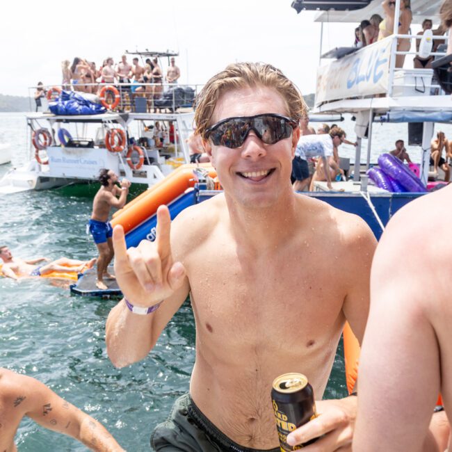 A man in sunglasses smiles and gestures with his hand while holding a can. He's in the water near a boat party with other people around. Several boats are seen in the background, and others are swimming and enjoying the water.