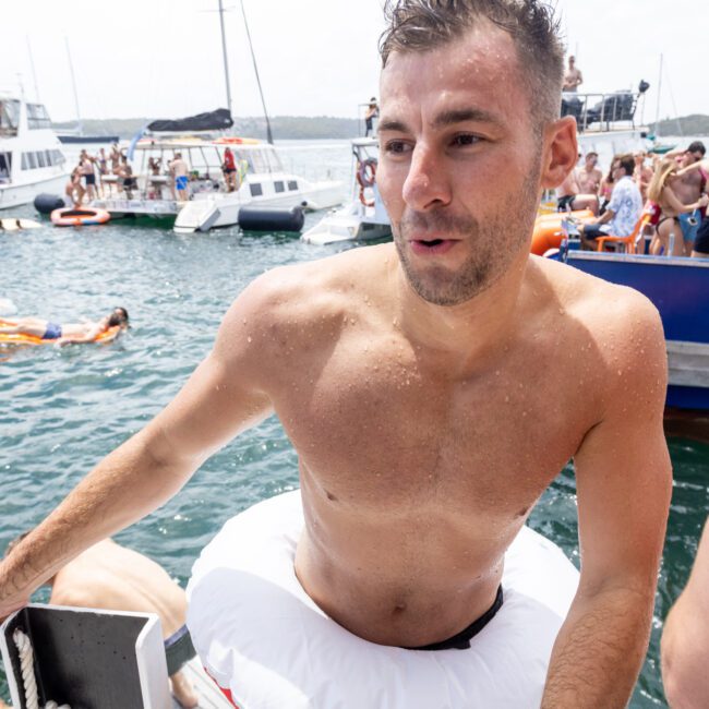 A man with a swimming ring climbs a boat ladder, surrounded by other people on boats and in the water. The scene is sunny and lively, with multiple boats and swimmers enjoying the day.