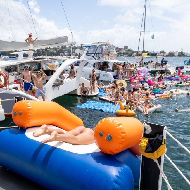 A crowded boat party on a sunny day, with people lounging on inflatables and swimming in the water. Boats are anchored nearby, and attendees enjoy the lively atmosphere, dressed in swimwear and surrounded by colorful floaties.