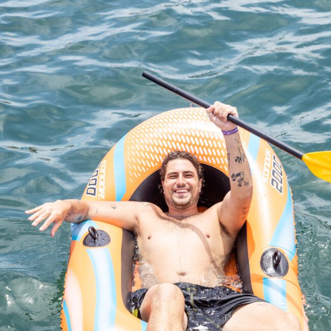 A man smiles while relaxing in an inflatable kayak on the water, holding a paddle. The kayak is orange and blue, and the water around it is calm and clear.