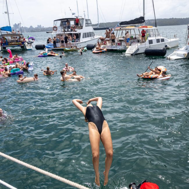 A person in a black swimsuit dives into the water from a boat, surrounded by people swimming and floating on inflatables. Several boats are anchored nearby, filled with people enjoying the sunny day.