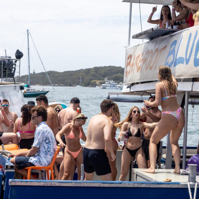 A group of people in swimsuits are on a boat party in a sunny bay. They are socializing and enjoying drinks. The banner on the boat reads "Barefoot Blue." Other boats and lush, green hills are visible in the background.