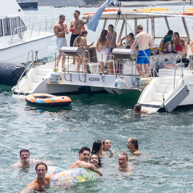 A group of people enjoying a sunny day on a boat and in the water. Some are aboard the catamaran, while others swim nearby. Inflatable floaties and smiling faces create a lively, relaxed atmosphere. Other boats are visible in the background.