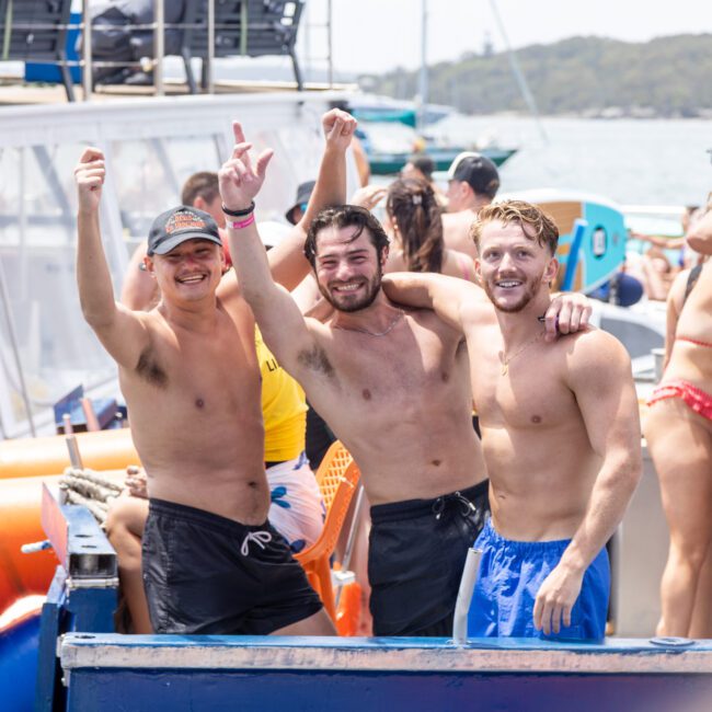 Three men in swim trunks standing on a boat, smiling and raising their arms in celebration. Other people are mingling around on the boat, with water and other boats visible in the background. The atmosphere seems lively and festive.