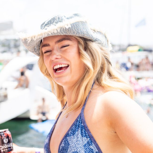 A woman in a blue bikini and denim bucket hat is laughing while holding a can of cider. She is outdoors at a sunny gathering on a boat, with other people and inflatables in the blurred background.