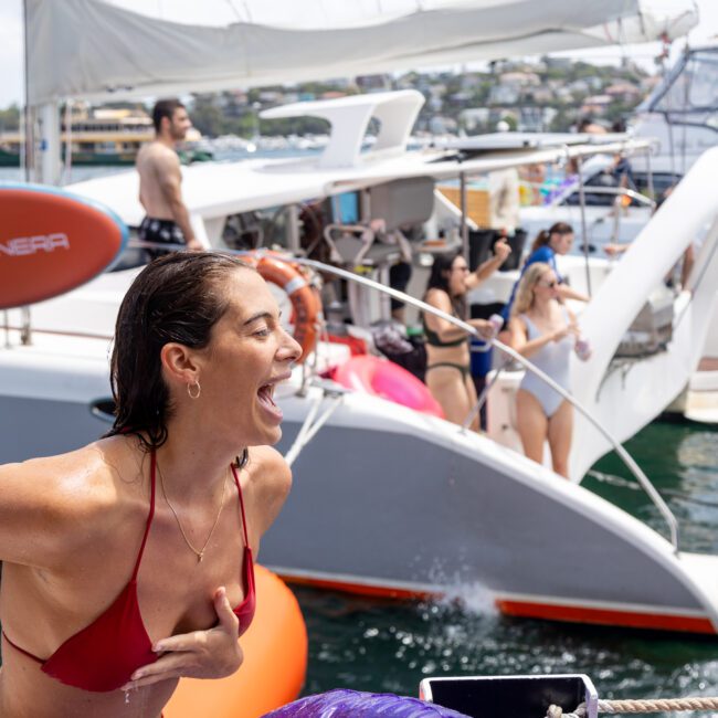 A woman in a red bikini laughs joyfully on a boat, surrounded by people enjoying a sunny day on the water. Other boats and swimmers are visible in the background, creating a lively, festive atmosphere.