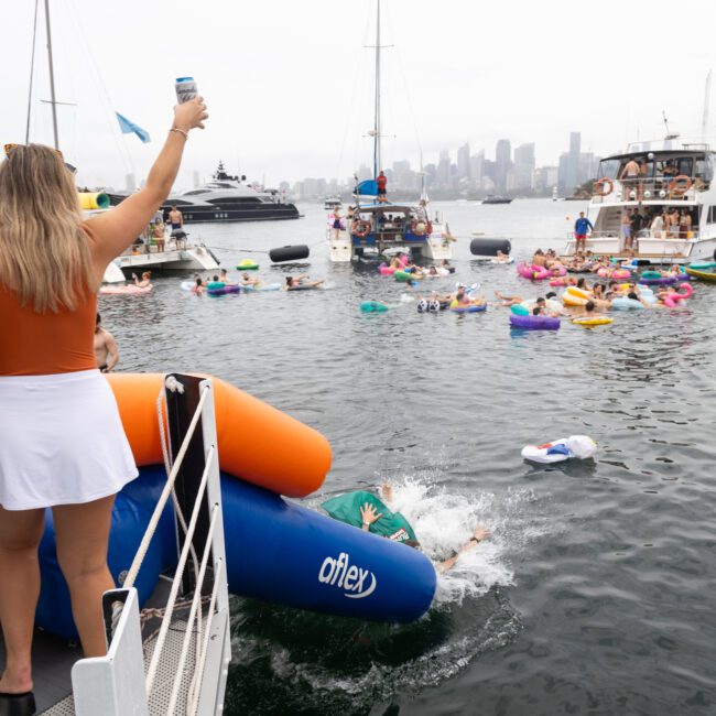 People celebrate at a waterside event with inflatable floats in the water. Boats are anchored nearby, and the skyline is visible in the background. A person stands on a platform, raising a drink in their hand.