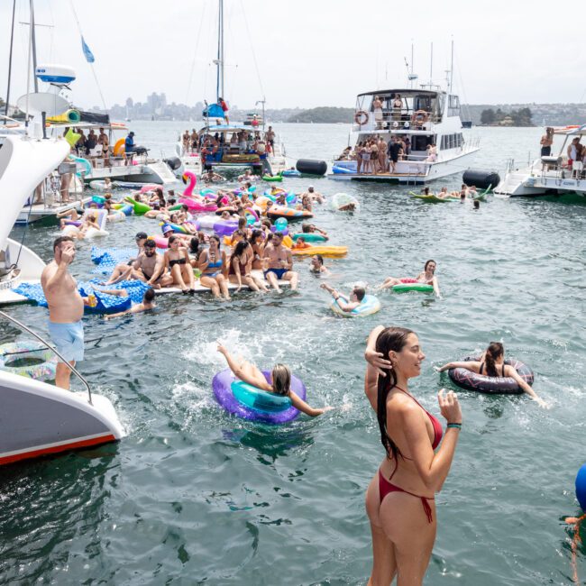 A woman in a red bikini stands on a boat deck, overlooking a lively scene of people on colorful inflatables in the water. Several yachts are anchored nearby, and the sky is clear. The setting is festive and summery.