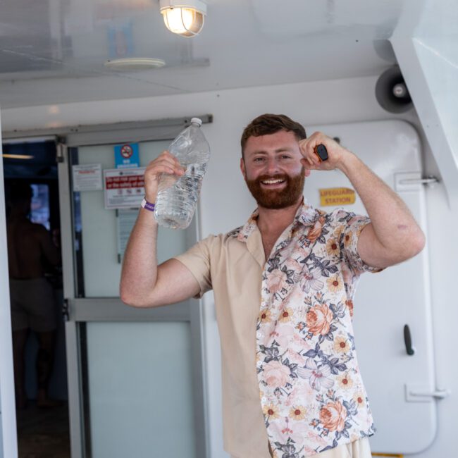 A man with a beard, wearing a floral shirt, smiles and holds a bottle of water and a black object on a boat. There are signs on the door behind him.