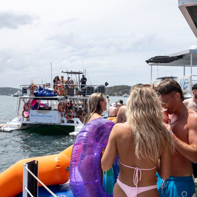 A group of people in swimsuits are enjoying a boat party on the water. One person holds a purple inflatable ring. Another boat with people onboard is in the background. The sky is cloudy, and the mood is lively.