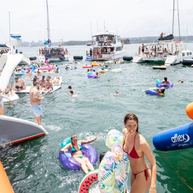 A woman in a red bikini stands on a boat's edge holding an inflatable. The water is crowded with people on various inflatables, and several boats surround the scene. The sky is overcast yet bright.