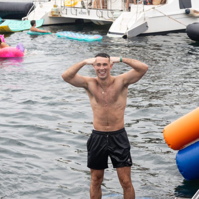 A man stands smiling on a floating platform in the water, with hands on his head. He's wearing black swim trunks. In the background, other people are swimming and relaxing near a boat. Colorful inflatables are visible around them.