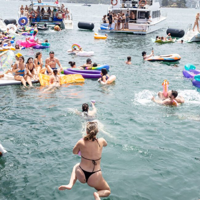 People enjoying a lively boat party in the water, with several individuals on colorful inflatable floats and one person jumping into the sea. A large boat is docked nearby, and the background includes a city skyline under a cloudy sky.