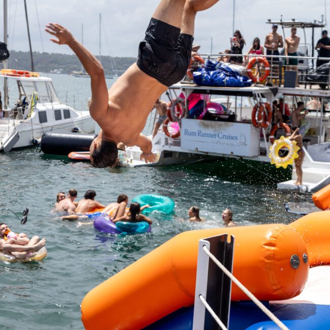 A man in swim trunks is mid-air, flipping off an inflatable water slide into the ocean. Several people are in the water with floaties. Boats are anchored nearby, and people are lounging on them under a cloudy sky.