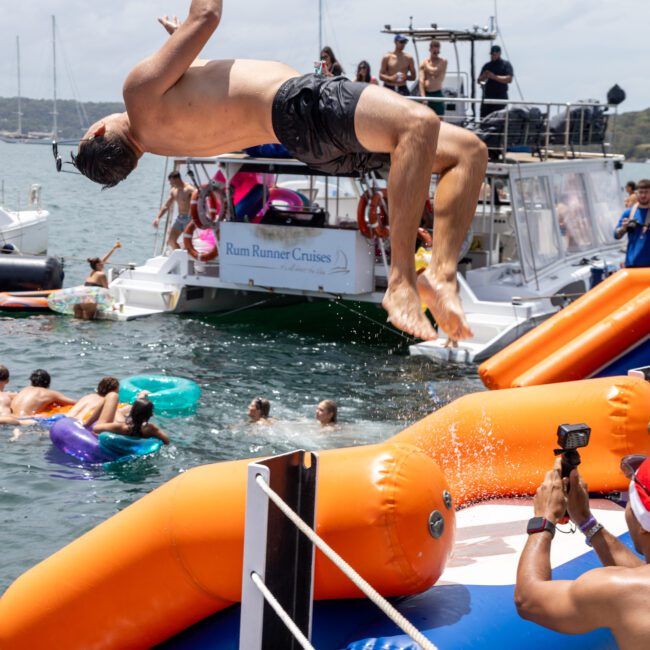 A person in swim trunks performs a backflip off an inflatable water slide into the ocean. Other people and boats, including "Rum Runner Cruises," can be seen in the background. A person is taking photos in the foreground.