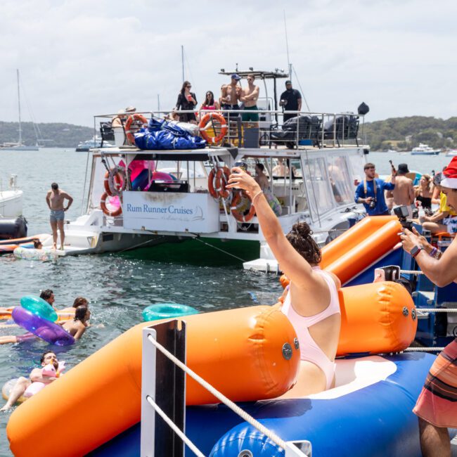 A lively scene of people enjoying a party on boats and in the water. Some are on inflatables, while others are mingling on the decks. One person is wearing a Santa hat, adding a festive touch. The background features a cloudy sky and distant shoreline.