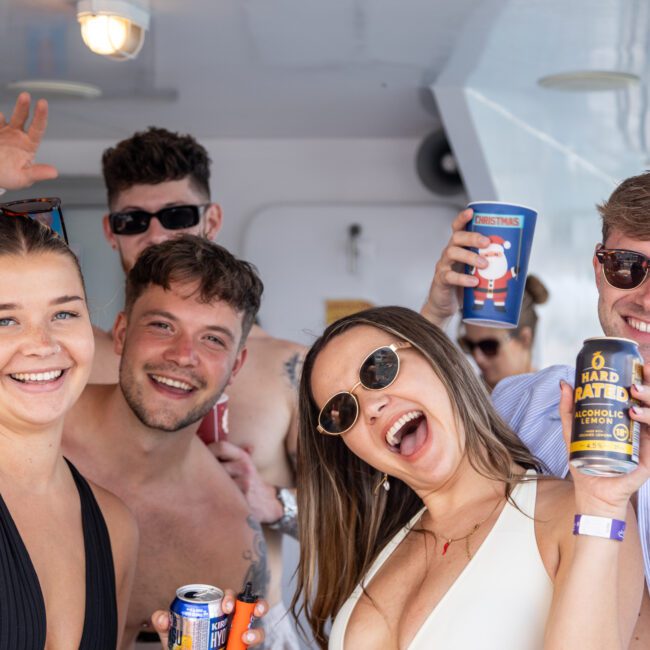 A group of five friends smiling and holding cans on a boat. They appear to be enjoying a sunny day with bright, cheerful expressions. Some are wearing sunglasses, and others are gesturing joyfully toward the camera.