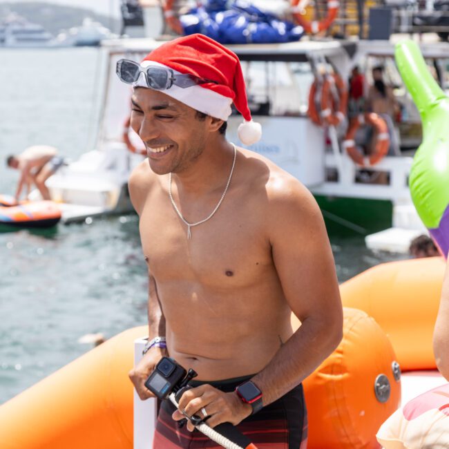 A person wearing a Santa hat and swimwear smiles on an inflatable water platform. They hold a camera and are surrounded by boats and people enjoying a sunny day on the water.