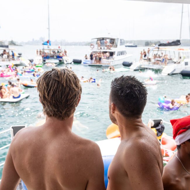 A large group of people enjoying a boat party on a sunny day. Many are in swimsuits, some in festive hats, with inflatable floats in the water. Boats are gathered together, and the atmosphere is lively and vibrant.