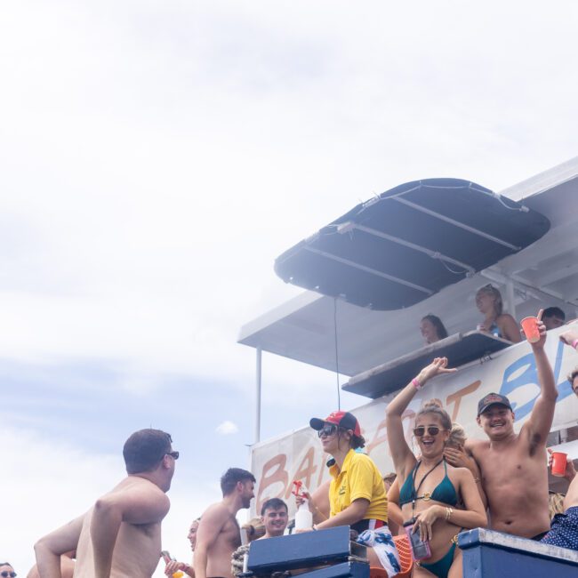 A group of people enjoy a boat party, with some in swimwear. One person stands behind a bar counter serving drinks. Others smile and wave, holding cups. The sky is bright, and there's a slide attached to the boat.