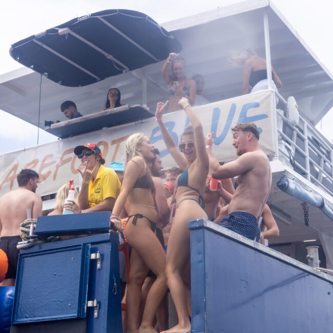 A group of people in swimwear are having a lively party on a boat. Some are dancing and holding drinks under a canopy, while others are enjoying the sun on a higher deck. A large banner is visible at the back of the boat.