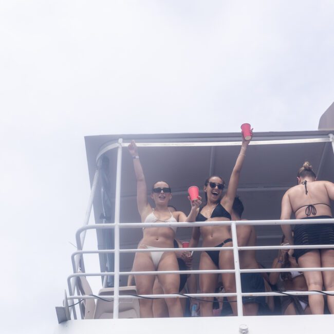 Two people in swimsuits stand on a boat deck, holding red cups and cheering. They are wearing sunglasses and appear to be enjoying a sunny day. Other people are in the background.