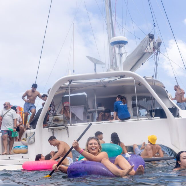 A group of people enjoying a day on a catamaran. Some are swimming or sitting on colorful floaties in the water, while others socialize on the boat. The sky is partly cloudy, creating a lively, summery atmosphere.