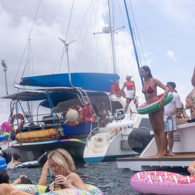 A crowded party scene on boats, with people in swimsuits enjoying the water. Some are on inflatable donuts and pool floats near catamarans under a cloudy sky. The atmosphere is lively and festive.