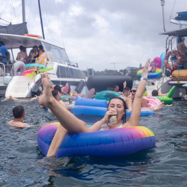 A person relaxes on a colorful inflatable ring, holding a drink, surrounded by others enjoying the water. Several boats and inflatables are in the background under a cloudy sky.