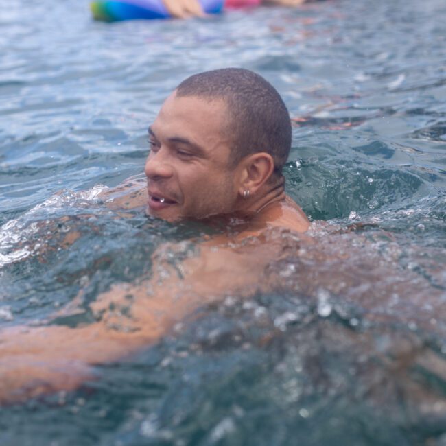 A person swimming in clear blue water with a colorful inflatable ring floating nearby. The swimmer appears joyful, and water splashes around them under a sunny sky.