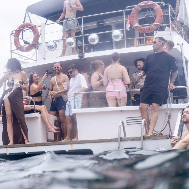 A group of people enjoying a party on a yacht. Some are on the deck taking pictures and talking, while others are swimming near the boat. The yacht is decorated with lifebuoys and silver balloons. The atmosphere is festive and lively.