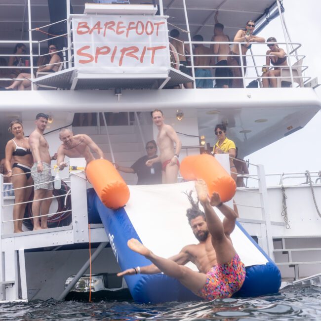 A group of people on a boat labeled "Barefoot Spirit." A man in colorful shorts is sliding down an inflatable slide into the water while others watch from the deck. The atmosphere is lively and festive.