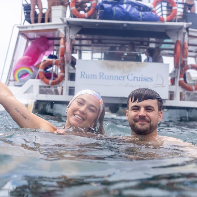 A smiling woman and man are in the water near a boat. The woman raises one arm joyfully. The boat's sign reads "Rum Runner Cruises." Several people and colorful inflatables are visible on the boat.