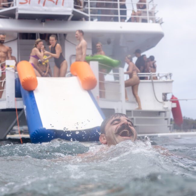 A man with a mustache swims joyfully near a yacht, surrounded by people enjoying a party. The yacht has a slide leading into the water. Others are on the deck, holding inflatable toys and socializing. The atmosphere is festive and lively.