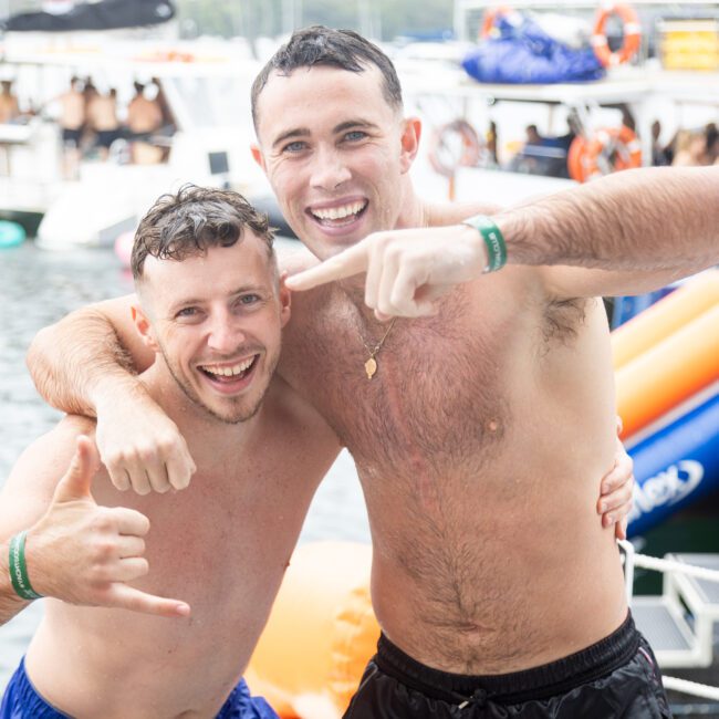 Two smiling men pose in swimsuits at a summer pool party, with one making a hand sign. People and floats are visible in the background, and a boat is docked nearby.