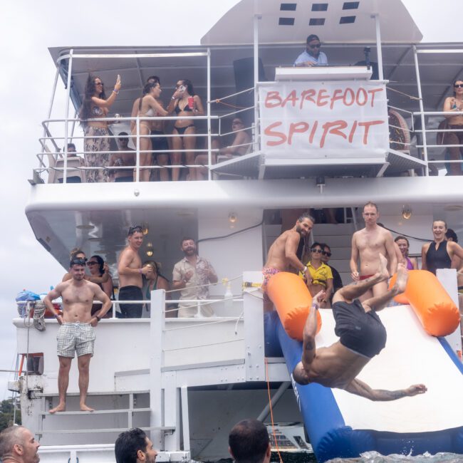 A group of people enjoying a party on a two-level boat with a "Barefoot Spirit" banner. A man is doing a backflip off an orange and blue slide into the water, while others watch and socialize on the deck. The sky is overcast.