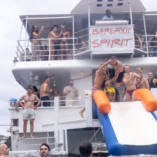 A group of people in swimwear are enjoying a party on a boat named "Barefoot Spirit." Some are using a slide to splash into the water. The atmosphere is lively and cheerful.