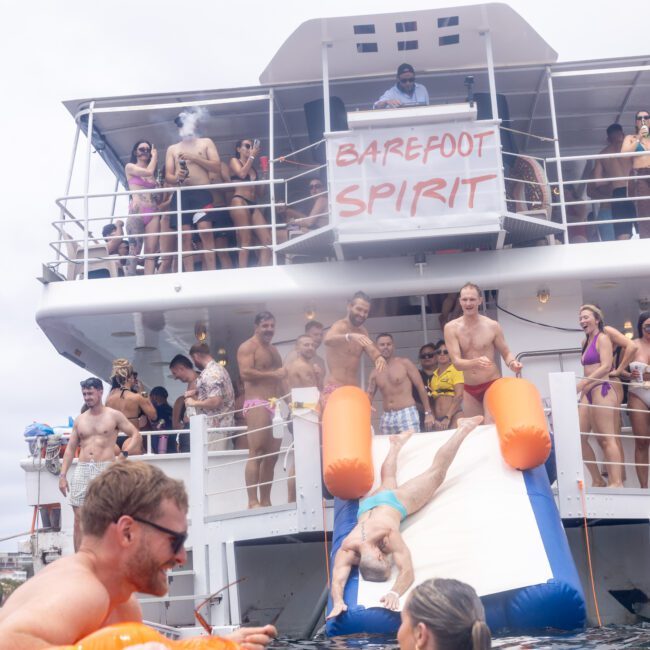 A group of people enjoying a party on a boat labeled "Barefoot Spirit." They are in swimsuits, with some on the deck and others using a slide into the water. The scene is festive and joyful, set against a cloudy sky.