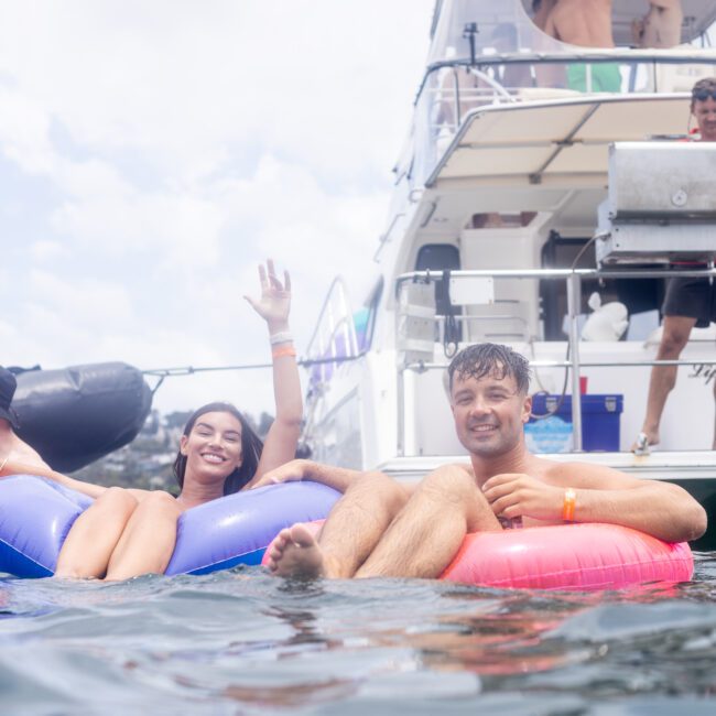 A man and a woman relax on inflatable rings in the water near a boat. The woman waves at the camera, and other people can be seen on the boat in the background. It's a sunny day with a partly cloudy sky.