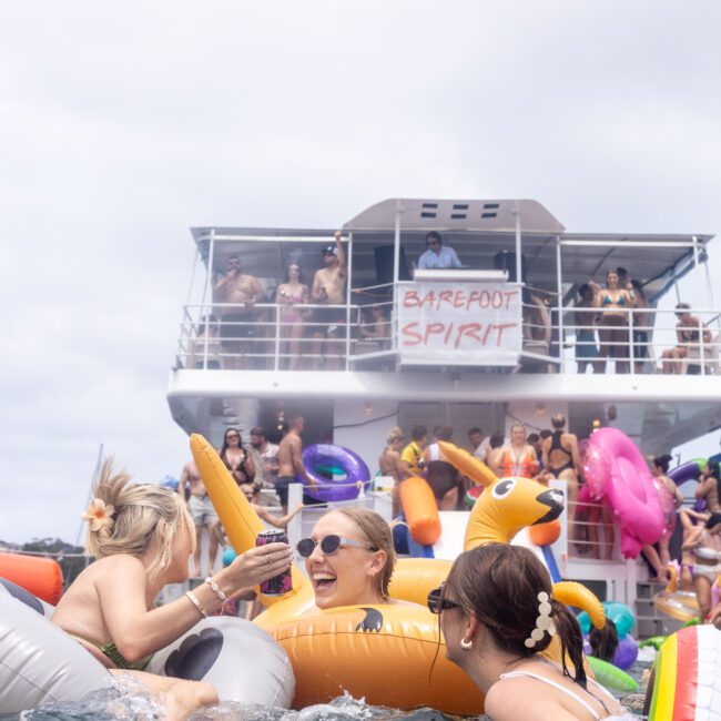 People enjoying a boat party with inflatable floaties. The boat's deck is crowded, and a sign on the boat says "Barefoot Spirit." The scene is lively, with people laughing and swimming in the water.