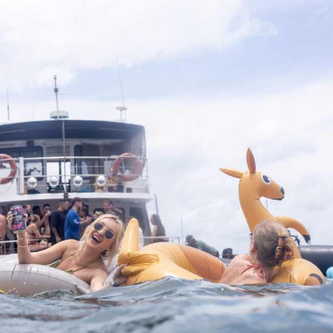 Three people enjoy a boat party, floating in the water on inflatable toys, including a kangaroo. A large boat is in the background with more people mingling on board.