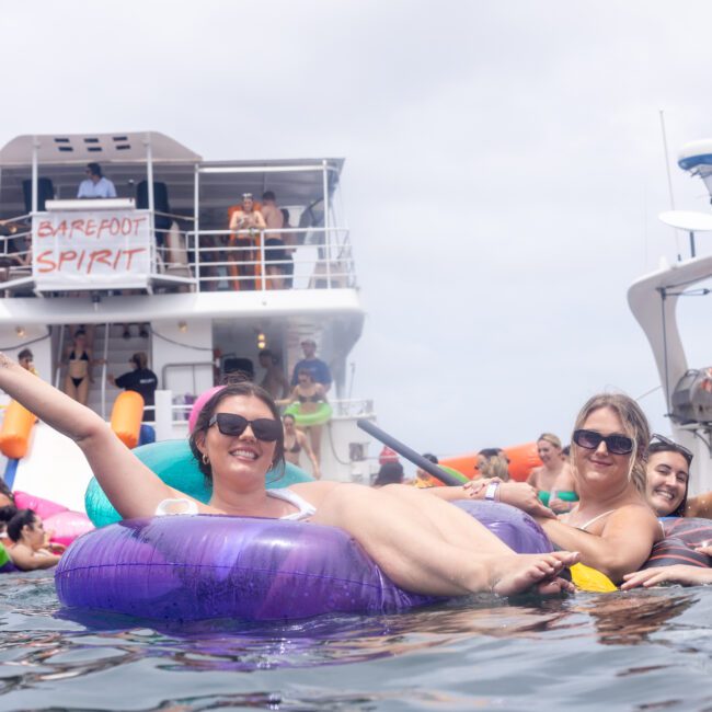 A group of people enjoying a sunny day on the water, floating on colorful inflatable rings near a large boat with "Barefoot Spirit" written on it. The atmosphere is lively, with many people on the boat and in the water.