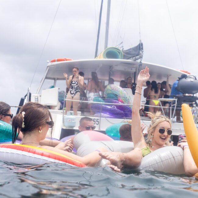 A group of people enjoying a sunny day on the water, lounging on colorful inflatables near a boat. Some are smiling and raising their arms in excitement. The background shows a cloudy sky and the boat anchored nearby.