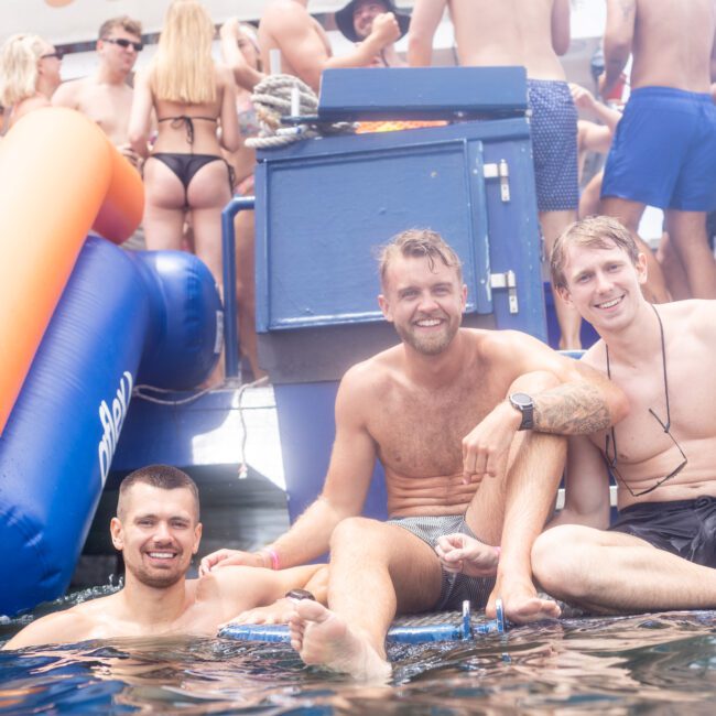 Three men are sitting on a raft in the water, smiling and posing for the camera. They are surrounded by a lively crowd of people in swimwear on a boat, suggesting a festive atmosphere.