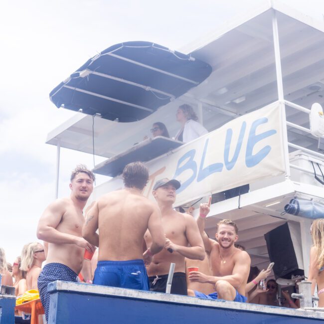 A group of people in swimwear enjoying a party on a boat called "BLUE." Some are standing and holding drinks, while others are sitting or leaning on the railing. The boat has two levels, with more people visible on the upper deck. It's a sunny day.