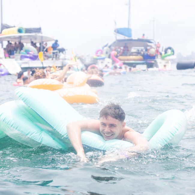 A person smiles while lying on a large inflatable float in the water, surrounded by others on similar floats and boats in the background, suggesting a lively and festive atmosphere.