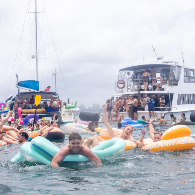 A group of people joyfully floating on inflatable rafts in the water, surrounded by yachts. The atmosphere is lively and festive, with many people enjoying the day under a cloudy sky.