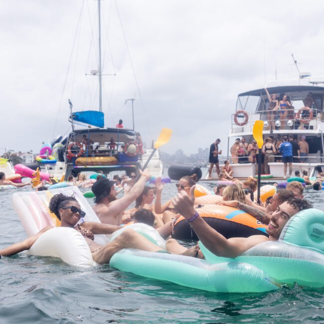 People are relaxing on inflatable rafts in the water, enjoying a sunny day. Boats are anchored nearby, and in the background, a cloudy sky is visible. Some individuals are holding paddles, creating a lively atmosphere.