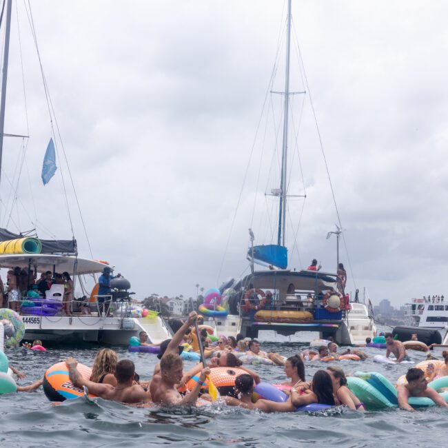 A large group of people relax on colorful inflatables in the water near several anchored boats. The sky is cloudy, and the atmosphere is lively and festive as people enjoy the day outdoors.