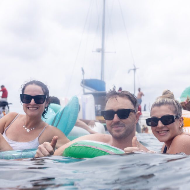 A group of people in sunglasses and swimsuits relax on colorful inflatable pool floats in the water, with sailboats in the background. The sky is cloudy.